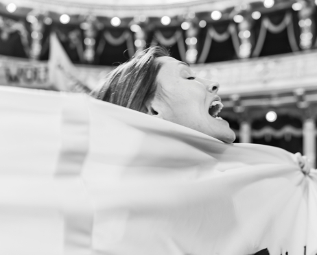 Czarno-białe zdjęcie. Kobieta stoi we wnętrzu teatru, jej głowa jest pochylona do tyłu, usta szeroko otwarte, jakby wykrzykiwała hasło. Przez pierwszy plan przebiega rozciągnięty materiał transparentu, który zasłania sylwetkę. W tle rozmyte rozświetlone balkony i dekoracyjne loże.